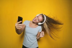 Young woman with headphones on is looking at her mobile phone and dancing, flicking her hair to one side. Mustard yellow backdrop.